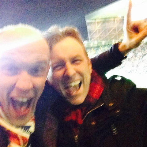 A blurry picture of two men celebrating in a stadium. They both wear scarves and jackets and have their arms around each other
