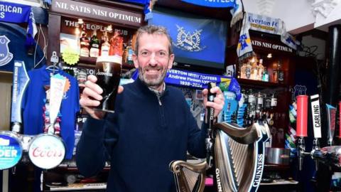 Dave Bond stands behind the bar at the pub holding a pint of Guinness. There are blue Everton scarfs and flags behind him.