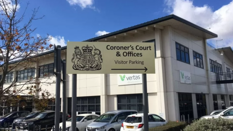 A view of Suffolk Coroner's Court from the outside. It shows the entrance sign with the building sat behind it. Several cars are pictured in the car park directly outside. 