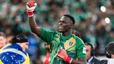 Edouard Mendy wears a green, short-sleeved Al-Ahli goalkeeping jersey and red gloves as he punches the air in celebration, while in the background a man is seen wrapped in a Brazilian flag and members of the media work wearing headphones