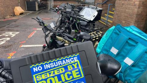 Electric bikes which were seized by police in Coventry. There is a large sticker with the words 'No Insurance' and 'Seized By Police'. A building and stairs can be seen in the background.