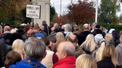 A crowd of dozens of people gather in front a large park area. A sign with "Hands Off Our Hospital" is visible in the background.