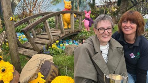 Two women sit in a highly-designed garden next to prop honey pots with a bridge in the background and model Winnie the Poo and Piglet characters.