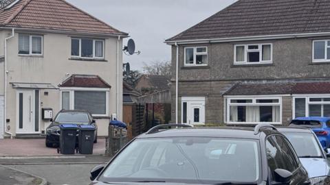 A view of the house from the street. It is a two-storey home with terracotta roof tiles and large driveway, with a white fence on the side of the house leading into the garden. There is another house made of grey brick on the right