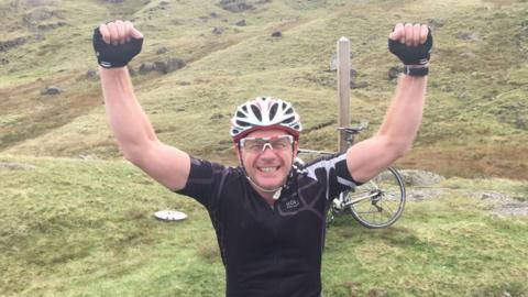 A man in a black cycling top and white cycling helmet with both arms held up in the air and a bicycle behind him on the grass