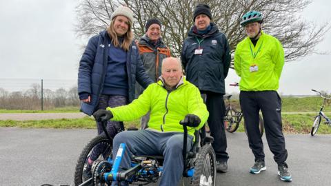 A man sits in a wheelchair trike wearing a high vis jacket. Around him stand 4 people smiling from the track. To the left is a young lady who wears a white hat and blue coat, then an older man who wears a black hat and light grey coat with orange detailing, next stands a man in another black hat wearing a dark grey coat and then stands a man in a high vis jacket and helmet.