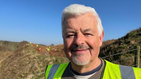 Win stood in front of one of the hillfort ramparts - huge earthen banks covered in grass and bracken. He's wearing a brown top with a yellow hi-vis vest over it. Over his right shoulder you can see about 10 volunteers - all also wearing orange or yellow hi-vis - working on the slopes of the hillfort. The sky is bright blue and clear, with strong sunshine. 