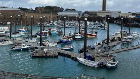 Boats and fishing boats in a harbour. A pontoon is floating on the service on the water which makes the boats accessible., The water is calm. Buildings are visible on land surrounding the harbour. The sky is grey.