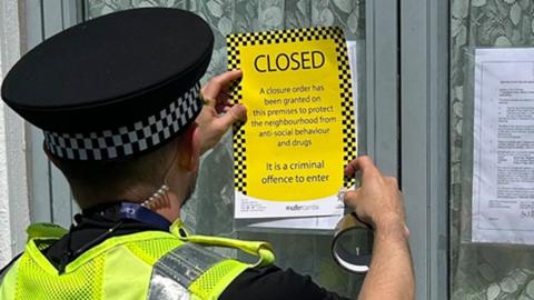 A male police officer is attaching a yellow "closed" notice to a front window of a house. The window has net curtains and another explanatory notice is attached to the other side of the window. The officer is wearing a police hat and a hi-vis yellow jacket with the word "police" across the back of it