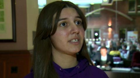 Emma, with long brown hair and wearing a purple hoody, looks to the side of the camera