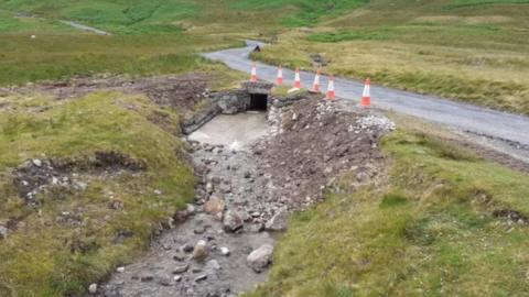 Cones on a section of a narrow mountain road at a culvert under which there is a rocky watercourse