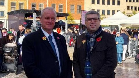 A man in a black wool coat, a white shirt and navy blue tie stands next to a man in a charcoal jakcet, and a navy tarten scarf. They are standing in front of several market stalls with lots of people waiting at them. An outdoor stage is set up behind them.