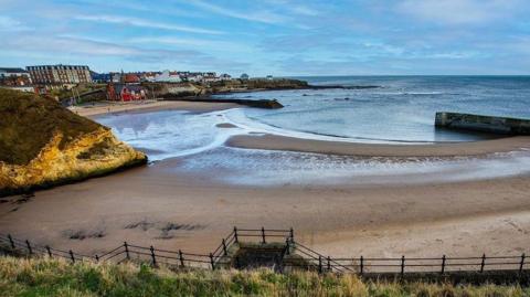 Cullercoats Bay. It shows sand and sea. There are houses and buildings in the distance.