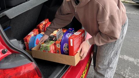 A car boot is being filled with hampers made from cardboard. The items include tea, crackers, chocolate and biscuits. The car is red and a young woman is lifting the hamper into the car.