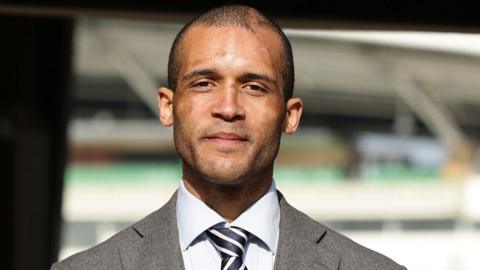 Head and shoulders image of Clarke Carlisle smiling with a sports ground blurred in the background. He is wearing a grey suit jacket, pale grey ad shirt navy and white striped shirt. He has very short dark hair and stubble with scars on his face and head