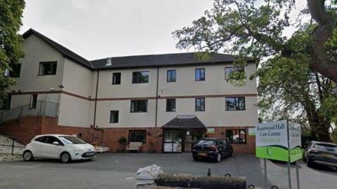 Image of the outside of a care home building, three floors high, mostly painted cream. Three cars are parked on the car park outside the building. To the right there is a white and green sign which reads 'Brentwood Hall Care Centre'. 