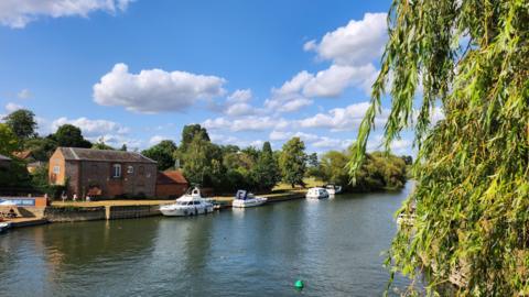 A river with three white boats moored on the far bank. There is a brick building and the river is flanked by green trees. Overheard the sky is blue with white clouds.