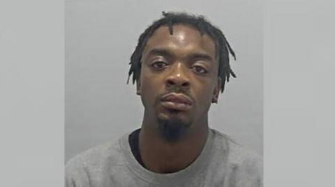 A police custody photo of Danderson Decker. He is looking at the camera, wearing a grey prison-issue sweatshirt. The background is also grey. 