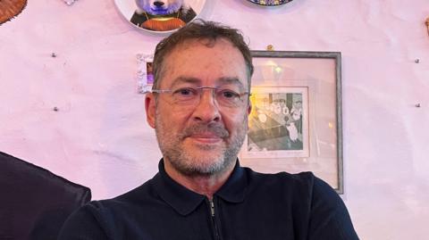 Simon Allum is a white middle-aged man with dark curly hair and grey stubble. He is wearing a black knit polo top and glasses with no frame. He is smiling at the camera with his mouth closed. The background is a pink wall with pictures and plates hung up on it.