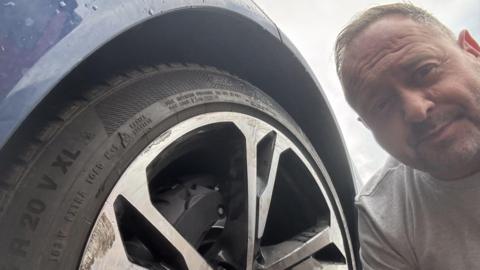 A man with short fair hair and a grey T-shirt crouches down by the wheel of his blue Audi. Although only part of the tyre is visible, the alloy wheel is scuffed.
