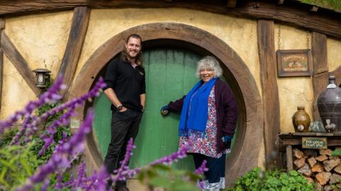 Miriam Margolyes and a guide stand outside a Hobbit house, with a circular green door and wooden beams