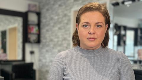 Sophie sits inside a modern hair salon, with styling stations, mirrors, and hair products visible behind them. The salon has grey décor and bright lighting.