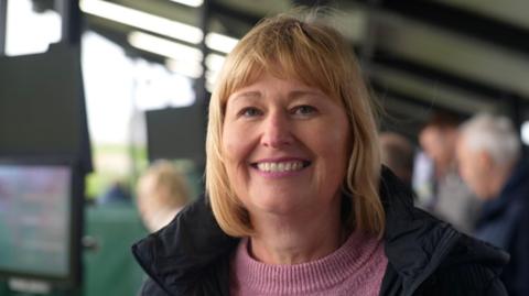 A woman with shoulder length blonde hair, wearing a pink knitted jumper and coat, standing outside smiling at the lens. There are people out of focus behind her.