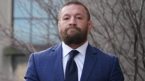 McGregor is wearing a navy suit with a white shirt and dark coloured tie. He has a thick brown beard and short brown hair. He has a straight face looking directly at the camera.