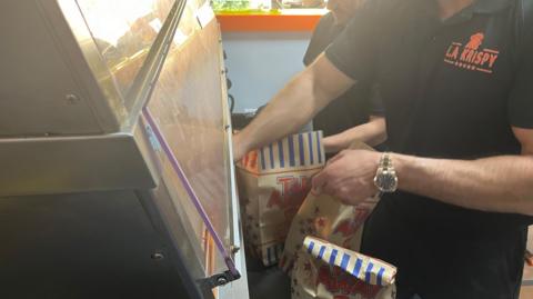 LA Krispy takeaway staff wearing black shirts with a logo in orange on them are packing orders into paper bags