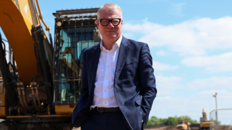 A man in a dark suit and white shirt is standing in front of a digger looking at the camera. He is wearing glasses.