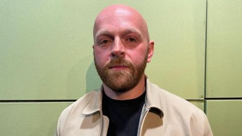 A bald man with beard stood against a light green studio wall. He is wearing a camel coloured jacket and black top