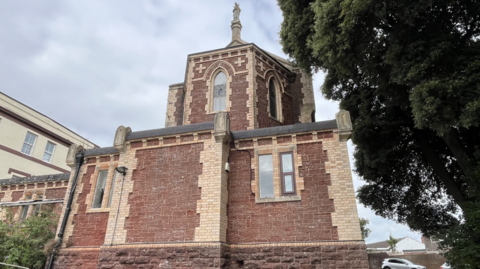 The image shows a historic red-brick and stone building with Gothic architectural features, including arched windows, ornate stone detailing, and a small tower topped by a statue.  The building is surrounded by trees and nearby structures, with a car visible in the background.
