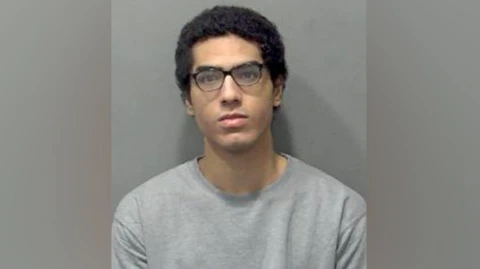 Custody image of Nicholas Prosper. He is wearing a grey sweatshirt and dark-framed glasses.