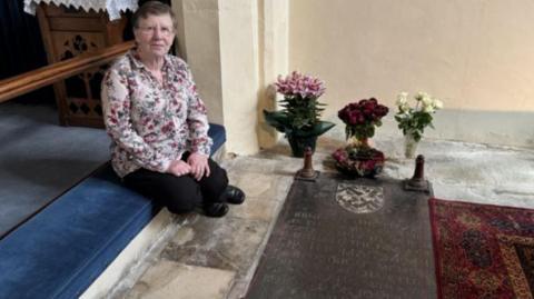 An elderly woman with glasses, short grey hair and floral shirt sits by the tomb of Sir Thomas More, which is covered by a large stone in the floor with inscriptions carved in it and flowers placed alongside   