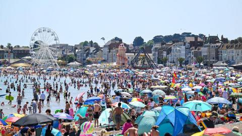 A general view of Weymouth beach during a heatwave in May, with hundreds of people using the beach and a blue sky above.