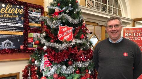A man with short grey hair and glasses wearing a grey Salvation Army jumper and blue shirt is smiling while standing next to a Christmas tree. The tree has tinsel and elves and a sign saying 'The Salvation Army'.