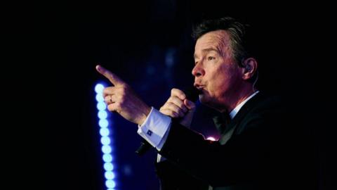 A man with dark hair and a dark suit in the dark, holding a microphone
