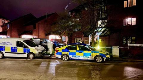 A number of police vehicles parked outside a block of flats at night. One van has the words FORENSIC INVESTIGATION on its side. A forensic officer in white overalls is visible from the road. There is also blue and white police tape across the entrance way to the block.
