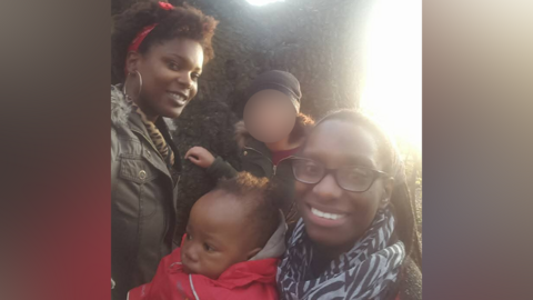 A young baby in a red coat and grey hood is in the middle of a family photo. A woman is holding him and wearing black glasses and a zebra-print scarf while smiling at the camera. A woman behind the baby wears a green coat and red scarf in her hair, while also smiling at the camera. Another figure behind the baby has their face blurred out.