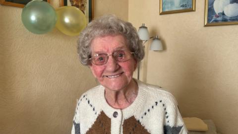 Woman with white hair, wearing glasses and a woollen cardigan, smiles at the camera. There are green and yellow balloons, a floor lamp and pictures on the wall behind her.