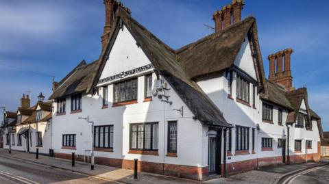 A large white thatched building is on the corner of a street and has a pavement which goes around the front of the building. The windows have red brick ledges, and there are three large chimneys. 