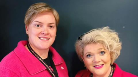 Two women in pink jackets smiling at the camera. They both have short blonde hair. The background is a blue grey colour. 