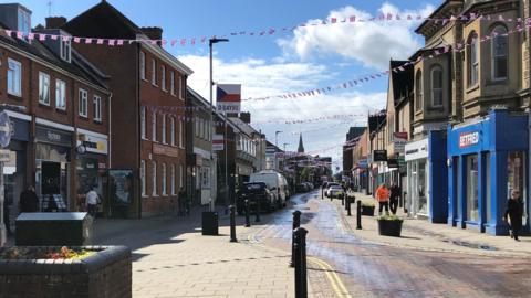 A town centre High Street on a sunny day. Bunting has been erected across the High Street between buildings. Cars are parked either side of a road. Some people are walking on the pavement toward the camera. Buildings line the side of the road and a church spire is in the distance.