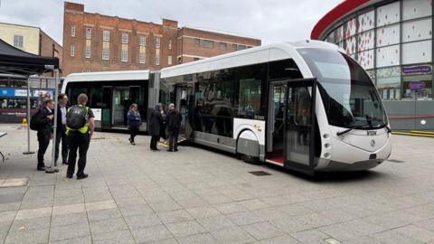 An articulated bus with a sloping front and concertina join between its front and back is on display in a town centre street.