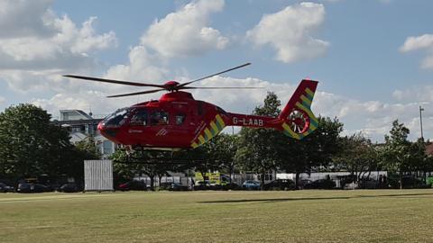 A red helicopter with yellow stripes on the tail hovers above the lawn of a large park, about 2 metres above it. You can see an ambulance on the road next to the park and cars parked alongside the fence. There are trees around the edge of the park.