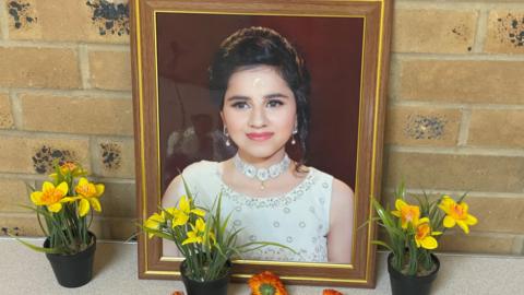 A photo frame with an image of a young woman with dark hair wearing a white dress and silver jewellery sits on a table, resting against a brick wall. Yellow daffodil plants sit in pots around the photo frame. 