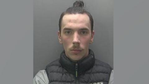 A slightly blurry police mugshot of David Gabryelski, who is clean shaven with a moustache and dark hair pulled into a bun on the top of his head. He is wearing a black puffa waistcoat over a grey top. 