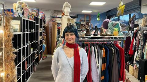 Ashira Zenab with black hair, light blue sweater and dark red scarf in the charity shop with a more contemporary look.