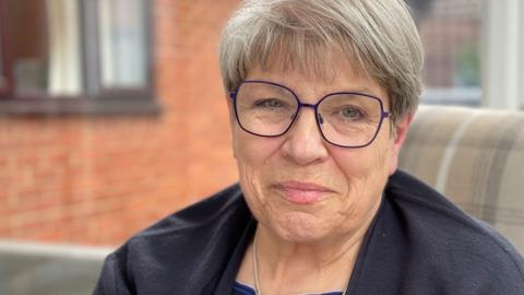 Diana Johnson, who has short grey hair, is wearing blue framed glasses and a black fleece. She sitting on a chair, with a window behind her and starting to smile. 