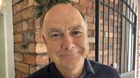 Kevin Allen smiling for the camera. He is a balding man who is clean shaven. There is a brick wall and a black gate behind him.